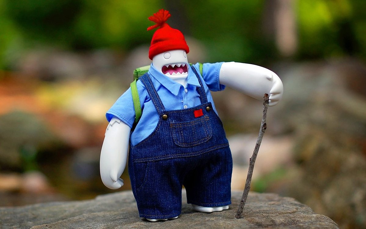 Bill the Yeti vinyl toy in a red knit hat, blue shirt and denim overalls, with a backpack and walking stick, standing on a rock outdoors.